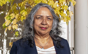 Photo of an Indigenous woman with shoulder-length, salt and pepper hair – and wearing a white blouse and blue blazer – stands outside in front of a Craftsman-style house. The sun is shining, and a climbing plant behind her has golden fall leaves.