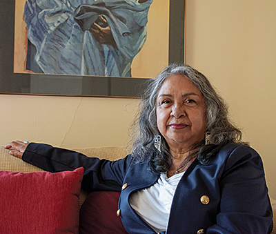 An Indigenous woman with shoulder-length, salt and pepper hair – and wearing a white blouse and blue blazer – sits on a red couch with her right arm stretched out, looking intently into the camera.