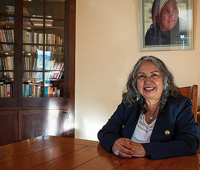 An Indigenous woman with shoulder-length, salt and pepper hair – and wearing a white blouse and blue blazer – sits at a wooden table with a glass bookcase to her right and a photo of an Indigenous elderly woman on the wall behind her.