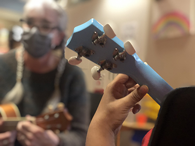 A close up of a child's hands on the fret of a ukulele with a woman with long gray hair in the background holding another ukulele.