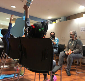 Two adults seated in chairs facing three kids with their backs to the camera. One kid has their hand in the air and and the other is holding up a powder-blue ukulele, demonstrating her fret positions.