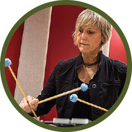 Circular photo with a forest-green frame of a white woman with short blonde hair playing the vibraphone. She's wearing all black, and you can see 3 of her 4 blue-headed mallets in the frame.