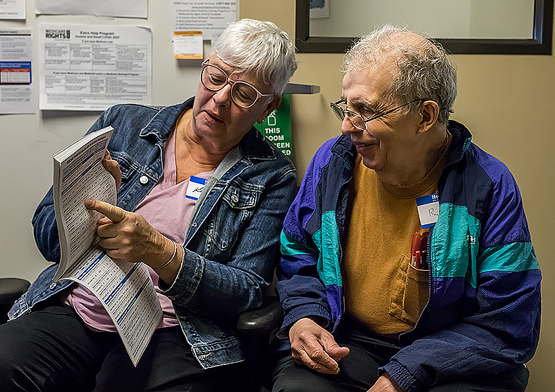 Two gray-haired adults talking to each other while holding and pointing to Medicare brochures.