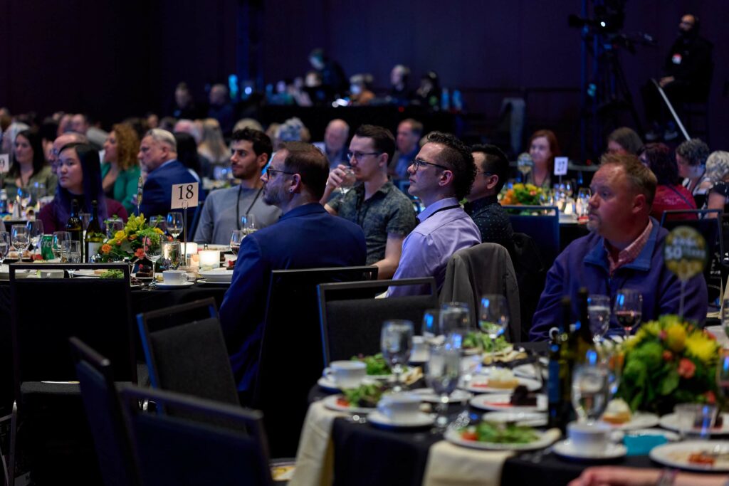 A crowd of people seated around tables with table clothes at a gala, all looking toward something out of the frame.