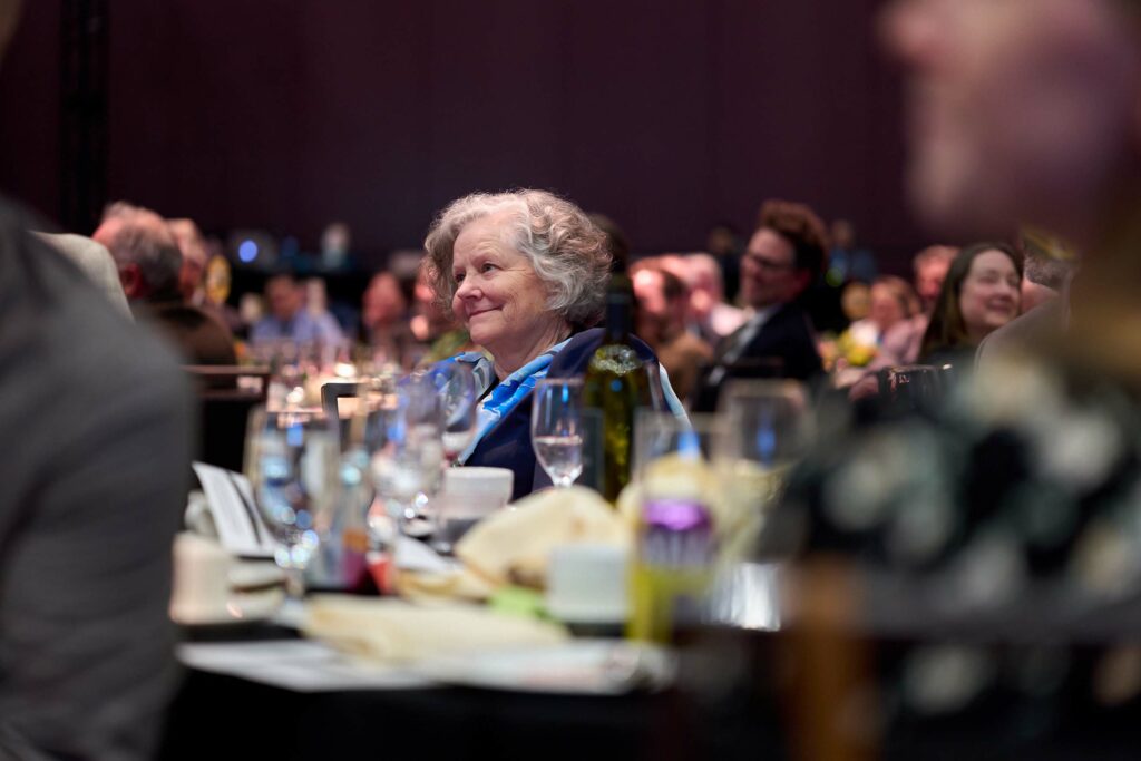 A shot of a crowd of well-dressed people at a gala, with one woman with gray hair in focus at the center.