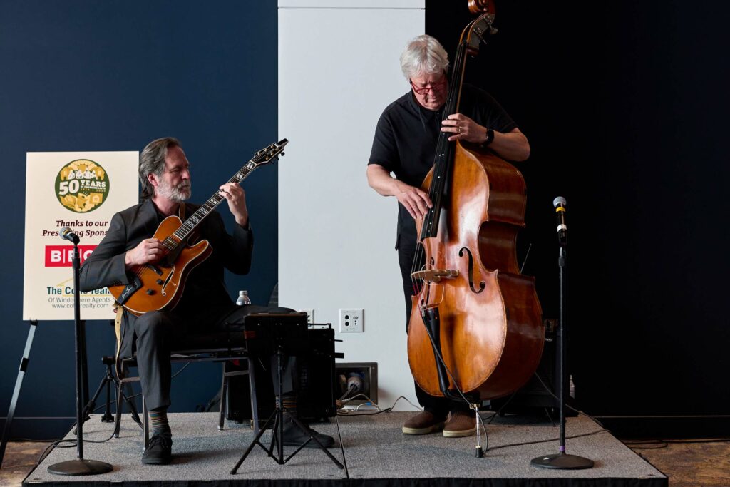 A seated man playing guitar next to another man standing up and playing upright bass.