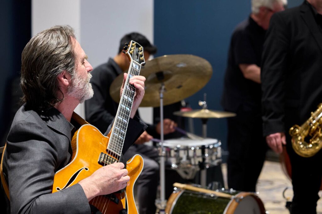 A man in a black shirt plays a guitar. A drummer with a drum kit is out of focus in the background.