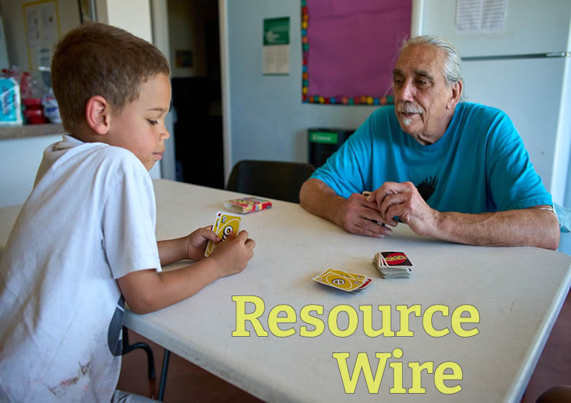 A boy in a white t-shirt examines a hand of Uno cards while sitting across a table from an older man wearing a blue t-shirt