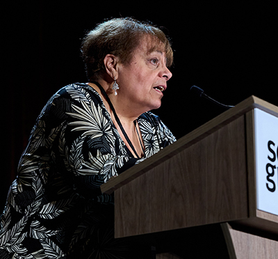 A woman in a black floral-print dress stands behind a podium that says "Solid Ground."