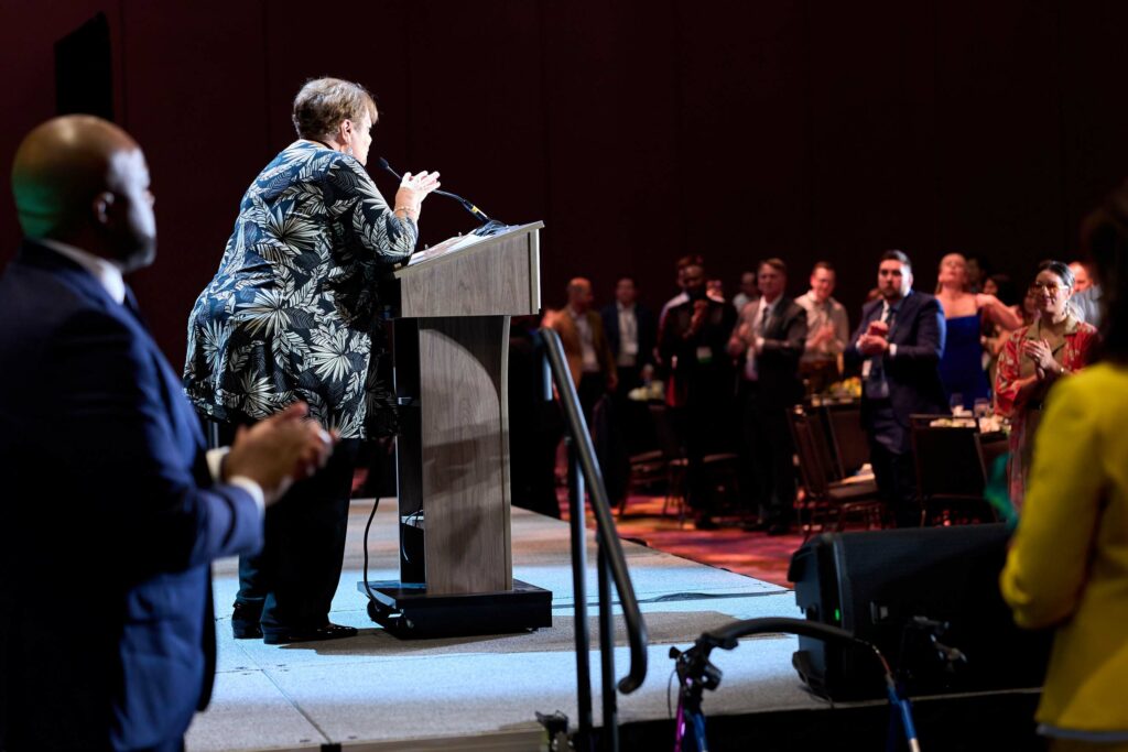 A woman in a black floral dress stands at a podium in front of a crowd.