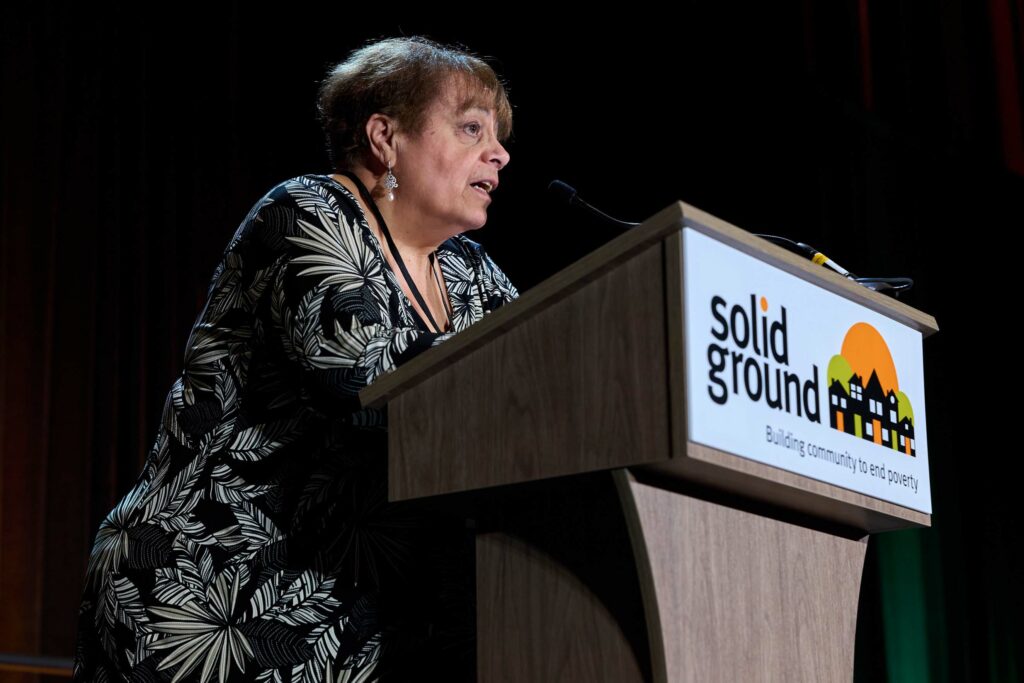 A woman in a black floral dress stands at a podium that reads "Solid Ground"