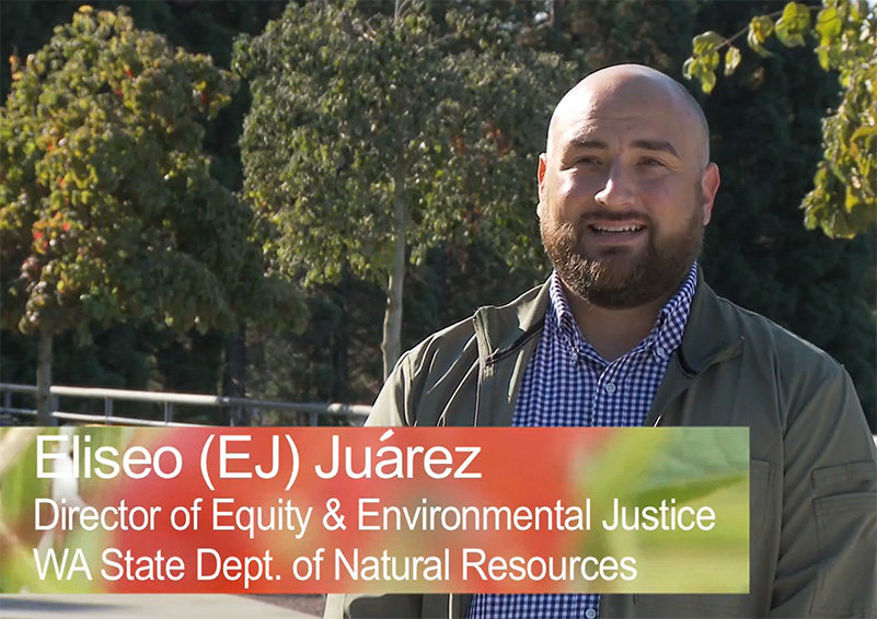 A screengrab from a video interview with a man in a green jacket and collared shirt in front of a stand of trees. At the bottom of the frame there's a banner that reads, "Eliseo (EJ) Juarez, Director of Equity & Environmental Justice, WA State Dept. of Natural Resources."