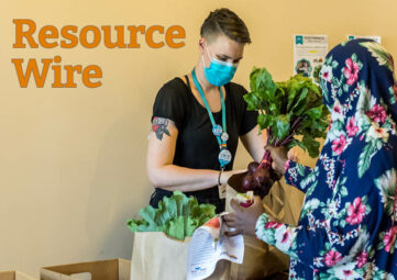 A women in a black t-shirt and medical mask stands across a table from a woman in a floral burka holding a bunch of beets over a paper bag. The words "Resource Wire" appear above them.