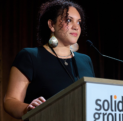 A woman with curly hair and long dangling earrings stands behind a podium that says "Solid Ground"