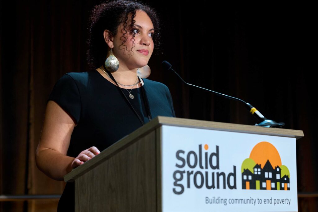 A woman with curly hair and long dangling earrings stands at a podium that says "Solid Ground."