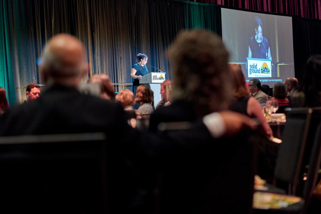 A woman with curly stands at a podium on stage, as seen from the back of a crowd that is out of focus in the foreground