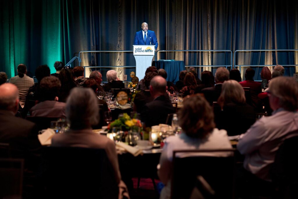 A man in a blue suit at a podium on a stage, seen from the back of a crowd at a gala.