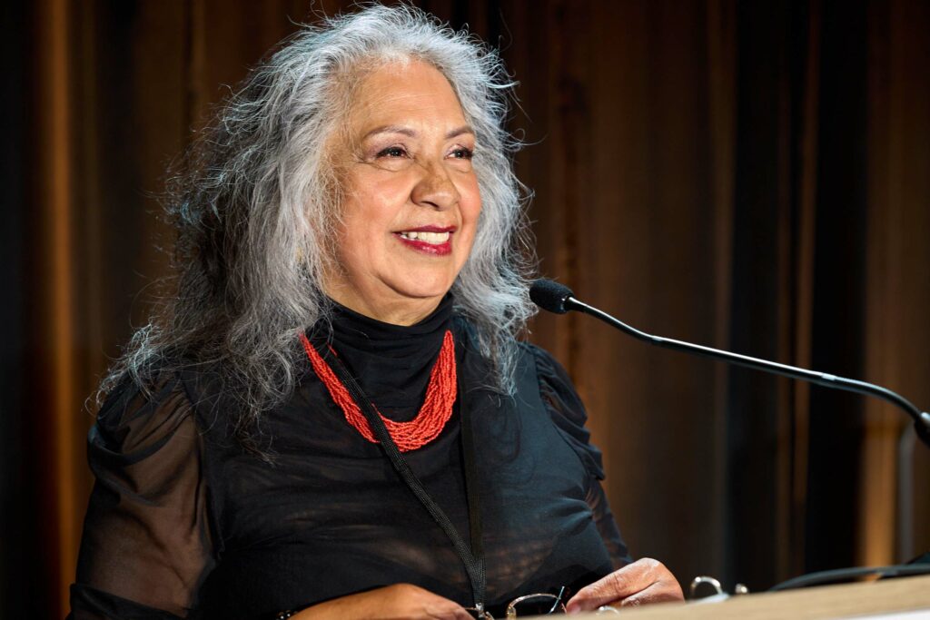 A woman with gray hair and a red necklace smiles as she speaks into a microphone.