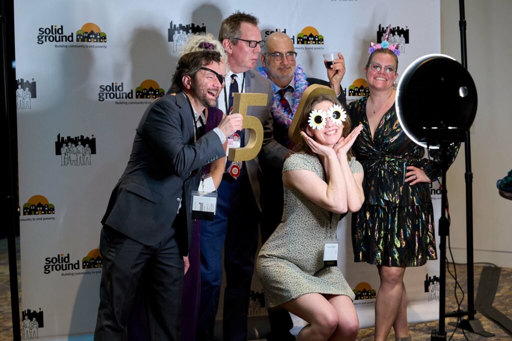 A group of people holding the numbers 5 and 0 make silly faces as they pose for a photo in front of a background with the Solid Ground logo.