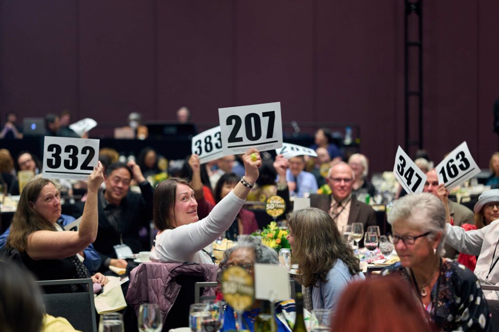 A group of people in a crowded ballroom hold up bid cards with different numbers.