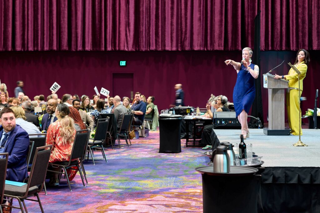 A woman in a blue dress on a stage points at one of many people seated around tables in a ballroom.