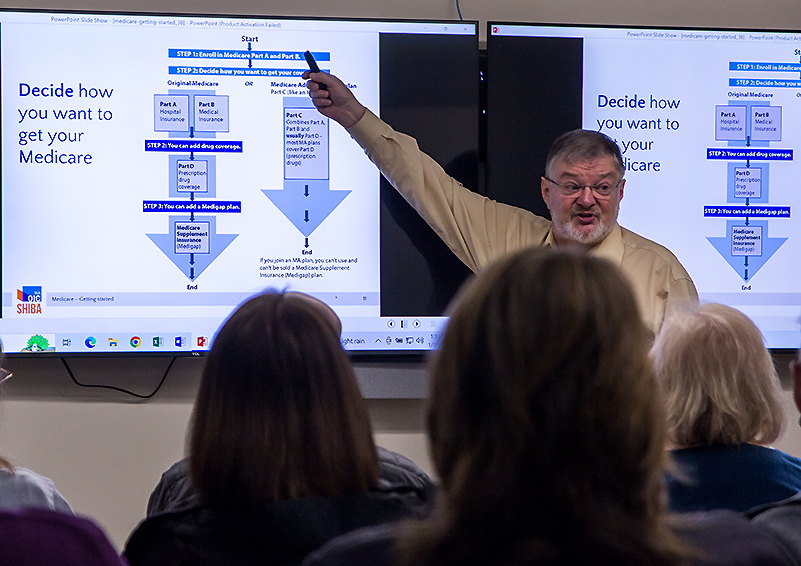 A man with grey hair and beard wearing glasses, an orange lanyard, and khaki pants and shirt, speaks to a small audience. He points to PowerPoint slides titled 'Decide how you want to get your Medicare.'