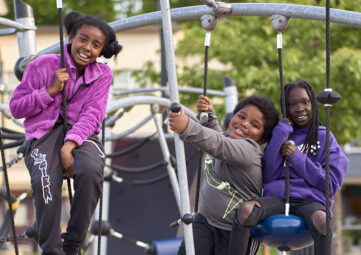 Three kids in jackets smile at the cameras as they hang from a climbing structure at a playground.