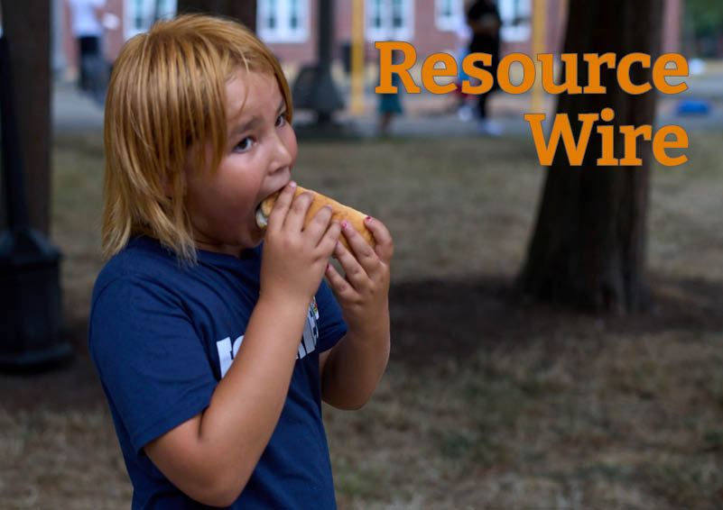 A boy in a park wearing a blue t-shirt takes a big bite of a hot dog. The words "Resource Wire" appear above him.
