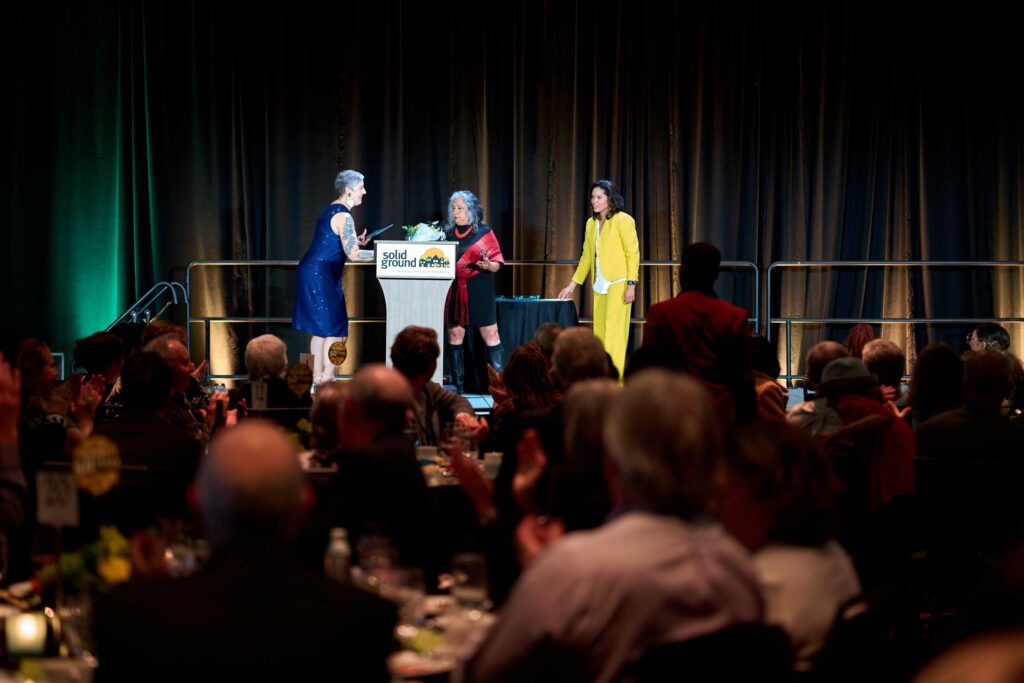 Three women stand on stage with a podium reading "Solid Ground" in front of a room filled with people at dinner tables.