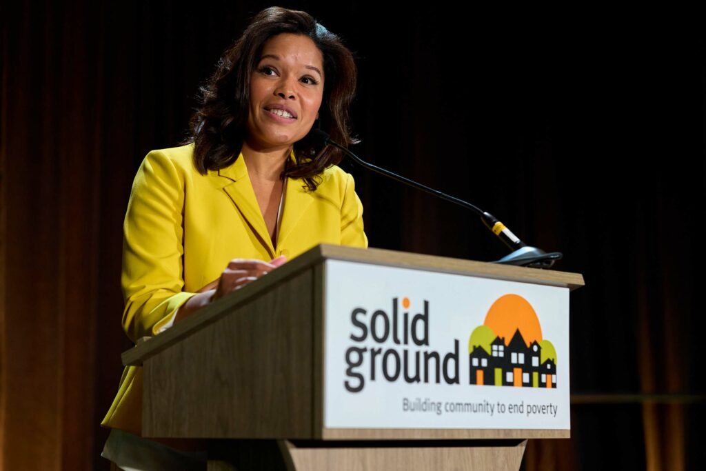 Woman with shoulder-length brown hair and wearing a yellow jacket speaks at a podium with the Solid Ground logo on it.