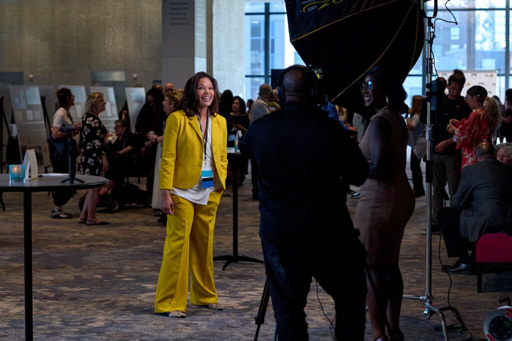 A woman in a yellow suit does an interview with a TV crew as a crowd mills about behind her.