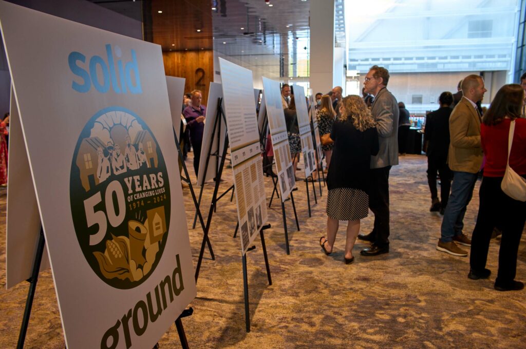 A couple examines one of several posters on easels. The closes one says "50 years" and "solid Ground"