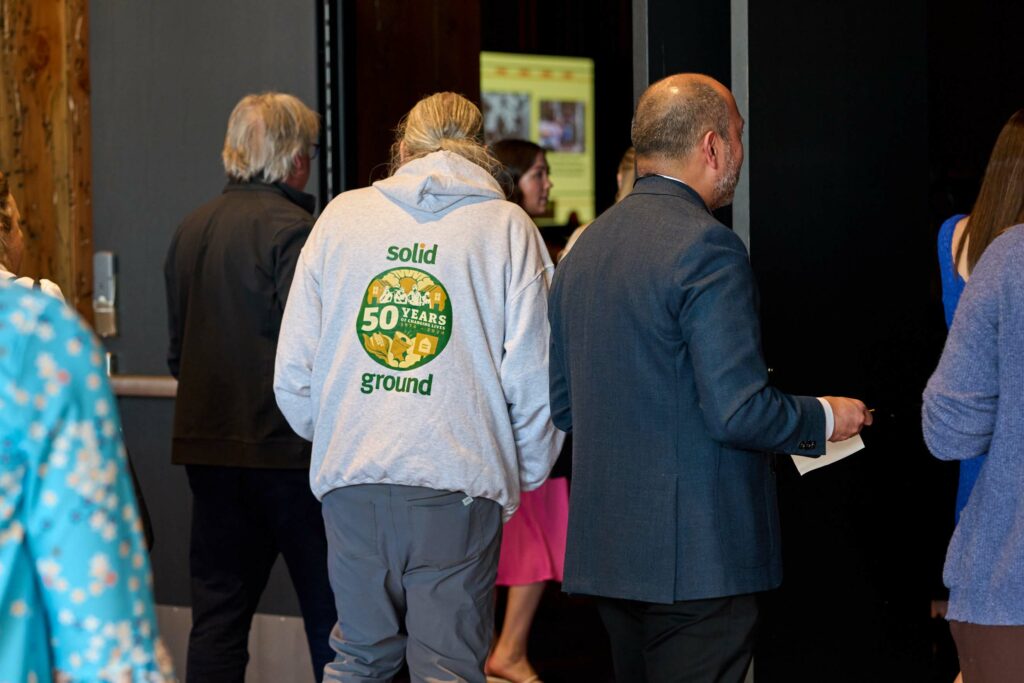 Several people walking into a room as seen from behind. One is wearing a sweatshirt that says "Solid Ground" on the back.