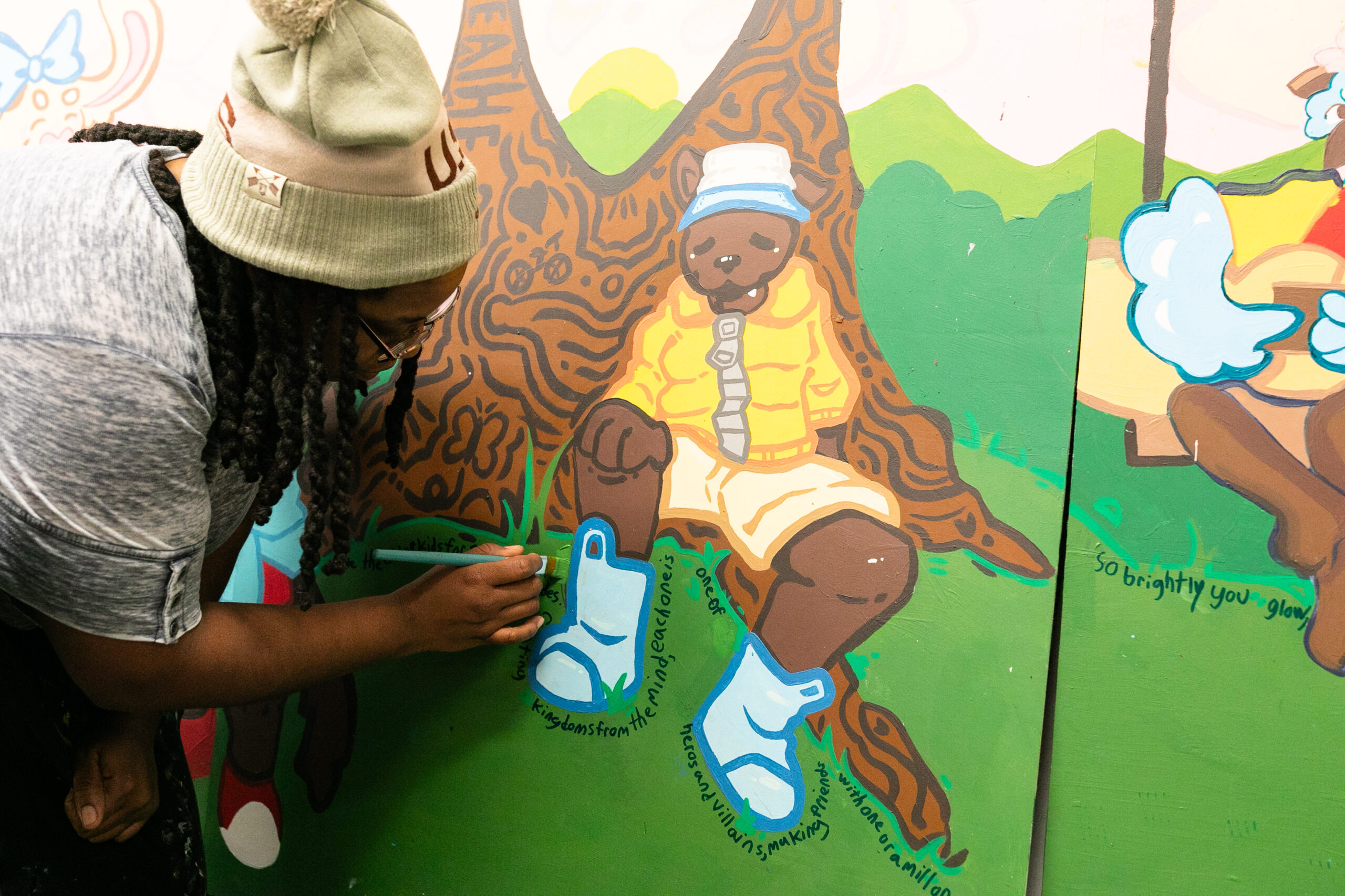 An artist brushes green paint on a part of a mural showing a cartoon animal seated at the base of a tree.