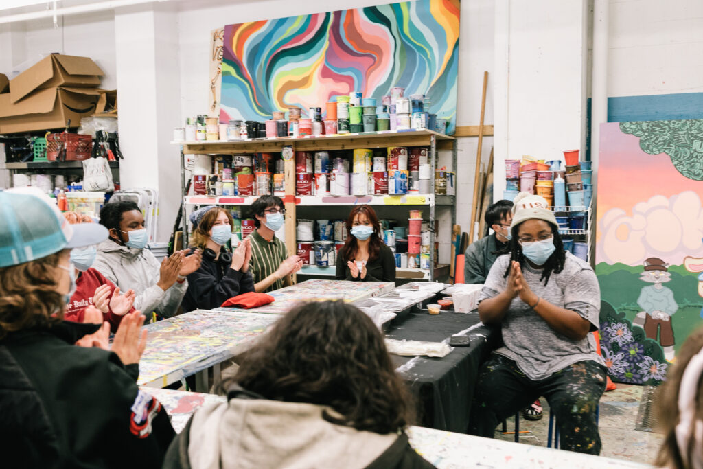 A group of young artists seated around tables in a studio clapping.