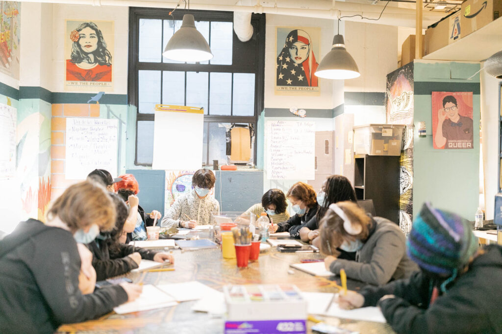 Around a dozen young people seated around a long table in a studio space drawing on pieces of paper.