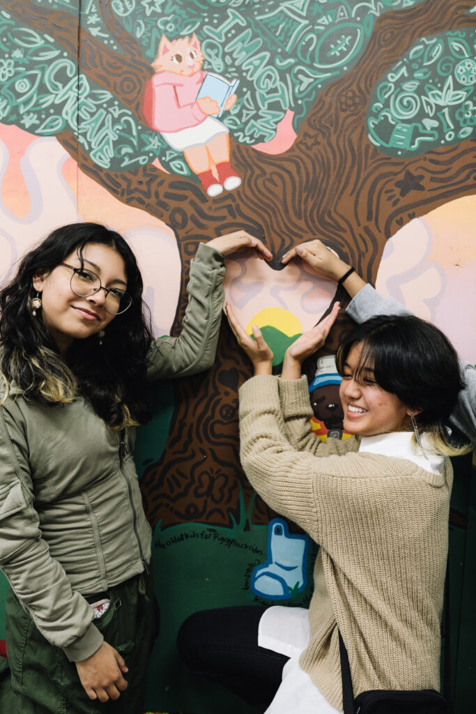 Two artists in front of the mural use their hands to make a heart shape over a heart-sharped hole in the trunk of a tree in the mural.