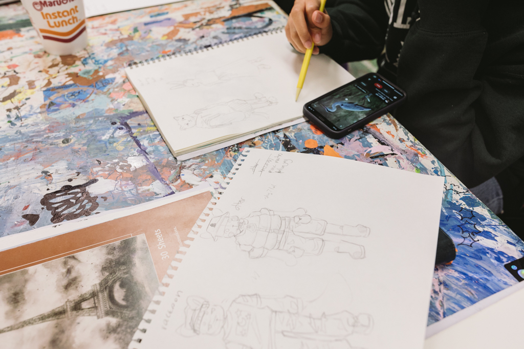 Close-up shot of an artist's hand and their sketches of animal characters for the mural, along with a phone showing a picture of a stork that the artist is using as a reference.