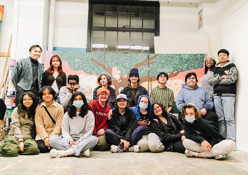 A large and diverse group of young artists sit and stand in a group in front a large, colorful mural in a studio space.