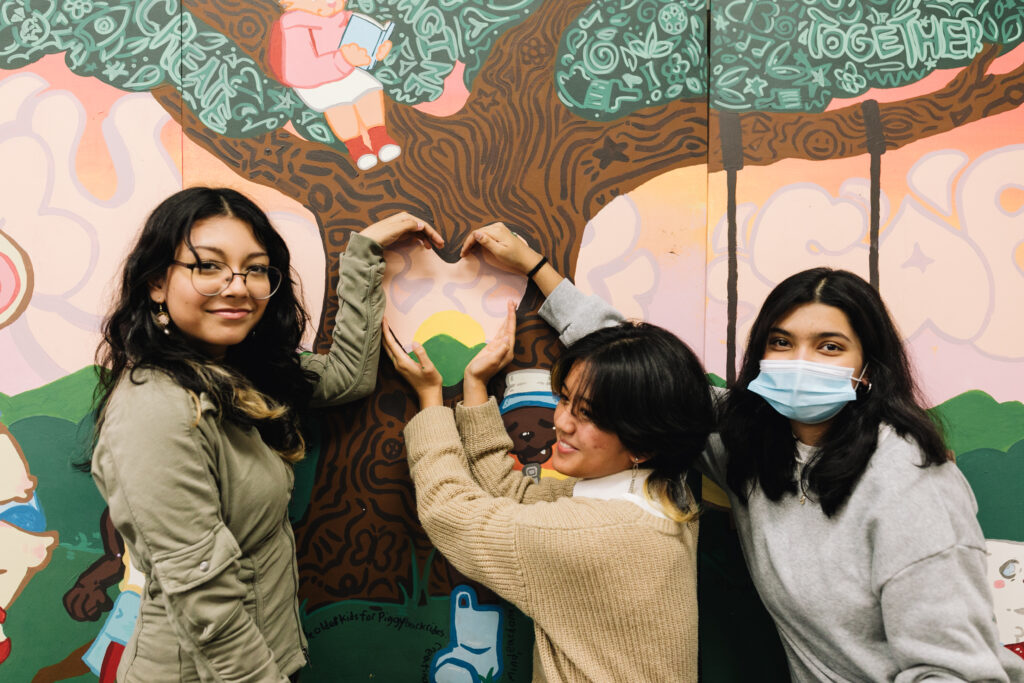 Three young artists in front of a mural use their hands to make a heart shape over a heart-shaped hole in the trunk of a tree in the mural.