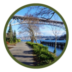 Circular photo with a dark green border of a paved path with a bridge span above it, trees and buildings to the left, and deep blue water and a pier to the right.