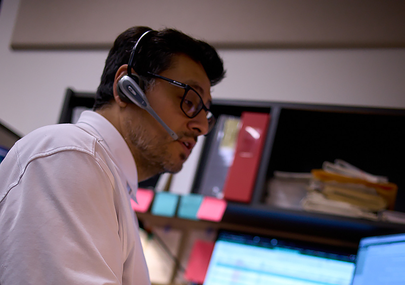 A man with dark hair and glasses talks with a headset on in front of a computer.