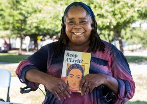 A smiling woman in a purple shirt holds a book in front of her titled "Keep A'Livin'"