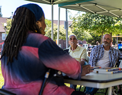 A woman in purple and blue with her back to the camera sits at a table and talks to a group of people in chairs under a tent.