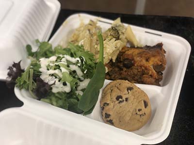 A food carton filled with casserole, pasta, chocolate chip cookies, and salad with dressing. 