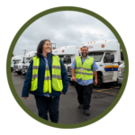 Circular photo with a dark green border of a smiling woman and man wearing safety vests and walking forward. Behind them are rows of paratransit buses.