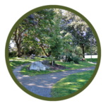 Circular photo with a dark green border of a gravel path winding through a park with trees and, a boulder, and a picnic table in the background.