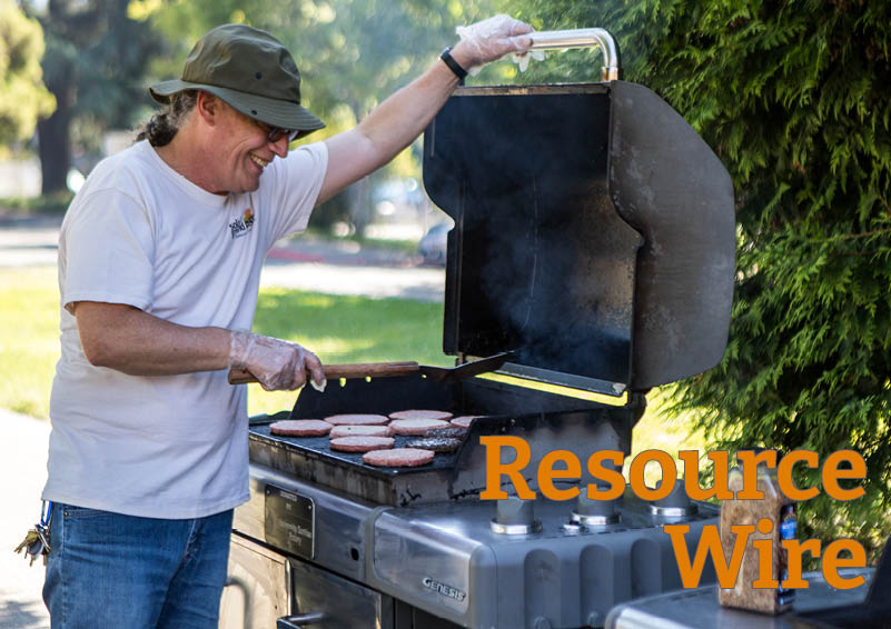 A man wearing a hat and a Solid Ground t-shirt flips hamburger patties while holding open the lid of a grill. The words "Resource Wire" appear at the bottom of the image.