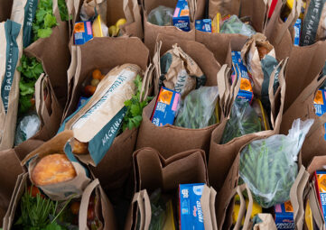 An overhead shot of brown paper bags filled with cereal boxes, baguettes, greens, and other groceries.