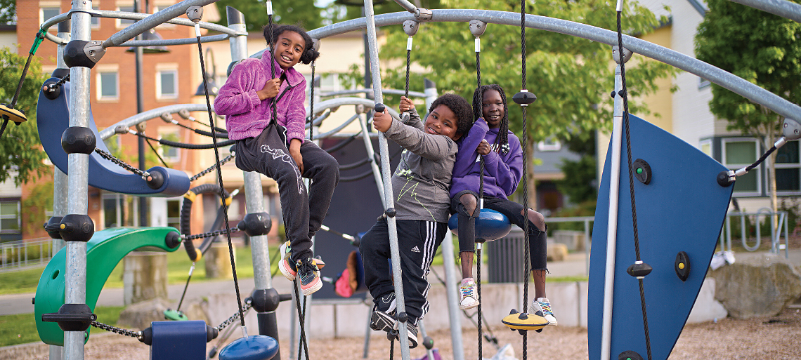 Three young Black kids sit on a jungle gym and smile into the camera. Behind them are trees and housing buildings.